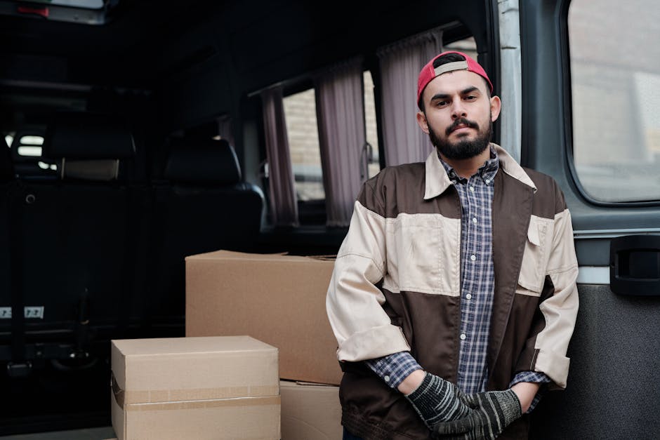 A man with a beard and wearing a blue beanie, a navy blue and red jacket, black pants, and white sneakers is sitting on the open rear door of a black delivery van. Inside the van, stacks of cardboard 