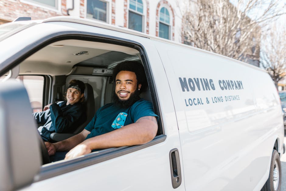 A young man with light brown hair, wearing a blue zip-up jacket over a white t-shirt and navy trousers, is sitting on the edge of an open-moving van with a neutral expression, holding a clipboard in h