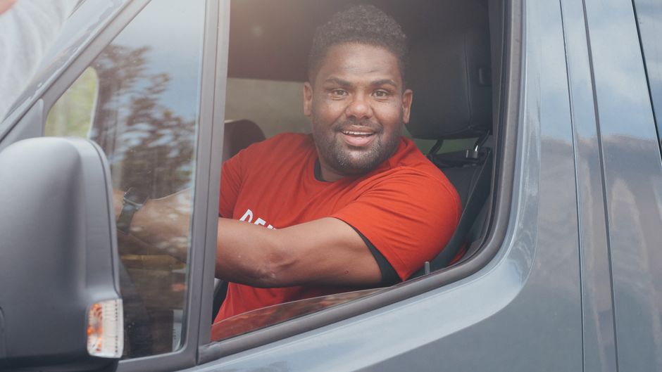 A man wearing a red cap, red T-shirt, and blue trousers is loading cardboard boxes into the open rear of a white van parked on a residential street. The van has its back doors wide open, revealing sev