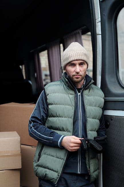A man wearing a white shirt and black cap standing outside a van loaded with multiple brown cardboard boxes of various sizes during a home relocation process. He is holding a large box with both hands