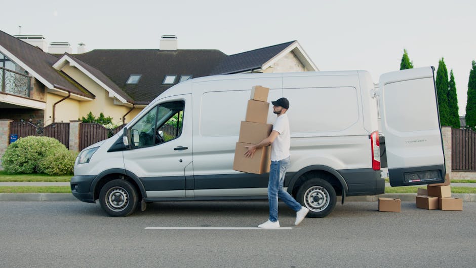 Two young women sitting on the edge of an open moving van’s cargo space, engaged in conversation and smiling, with cardboard boxes and wrapped items visible behind them. The woman on the left has cu