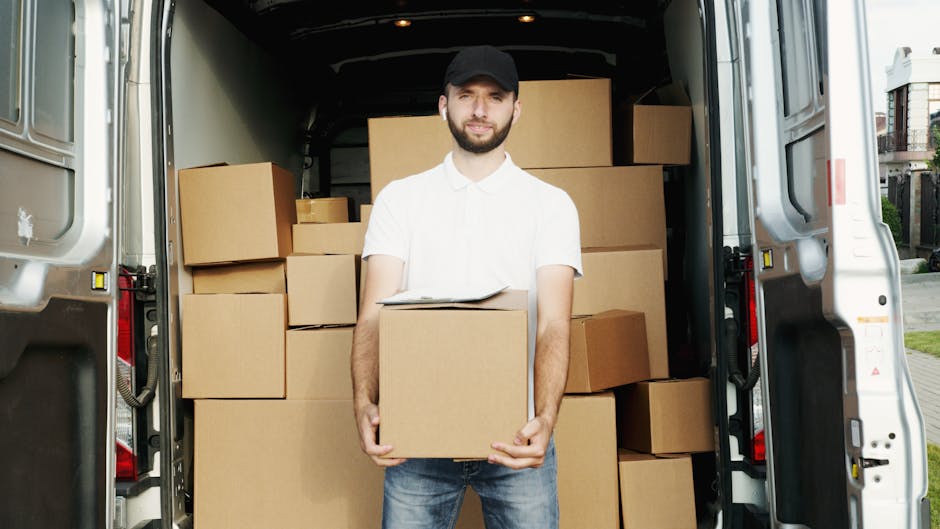 A man dressed in a light grey knitted beanie, a dark navy tracksuit, and a green padded vest standing next to a black van used for home relocation. Inside the van, partially visible behind him, are br